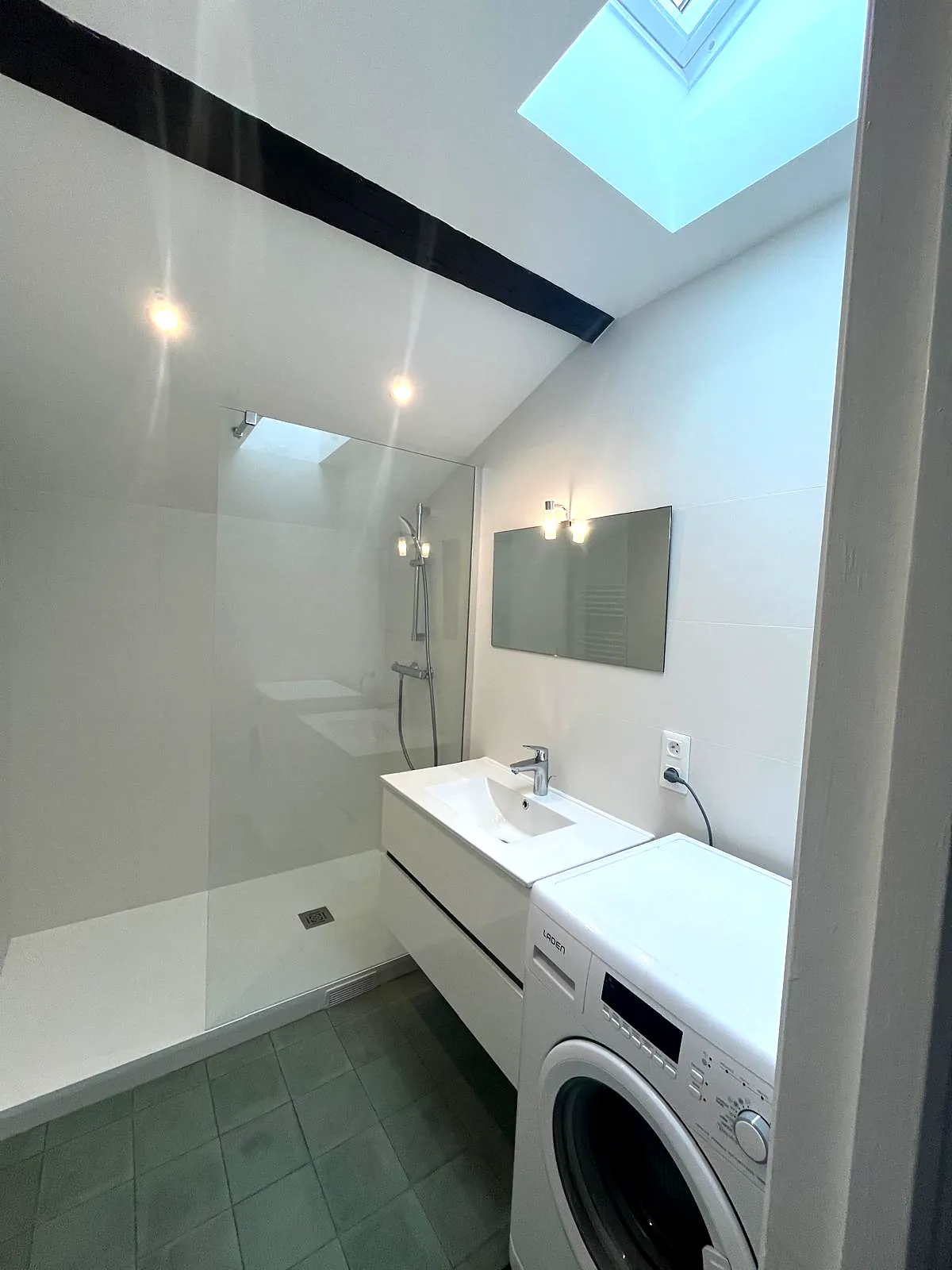 Salle d'eau terminée : douche, vasque et lave-linge sous la fenêtre de toit