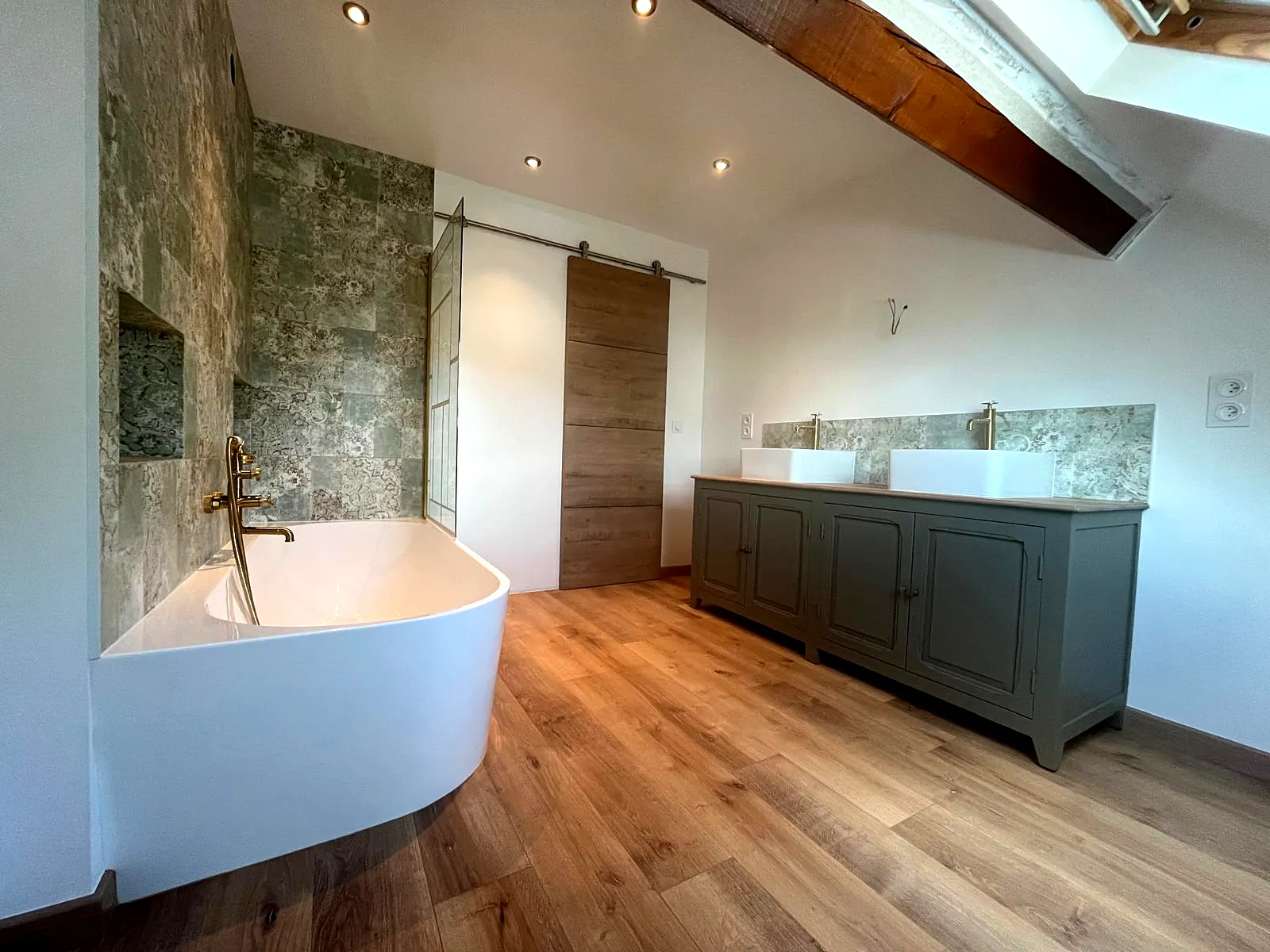 Suite parentale aménagée sous combles : baignoire îlot, mur effet pierre, sol effet bois et porte coulissante